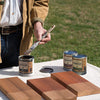 A man holding a stain brush with three different pints of Benjamin Moore exterior stain. 