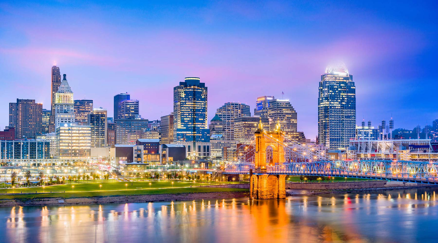 Cincinnati, Ohio skyline in the evening, showing a blue and purple colorful sky, with the lights from buildings reflecting on the water.